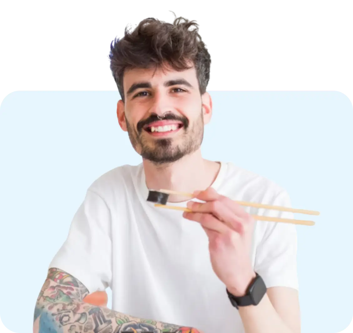 Smiling man with curly hair and beard holding chopsticks with sushi, wearing white t-shirt against light blue background, expressing joy while enjoying healthy food